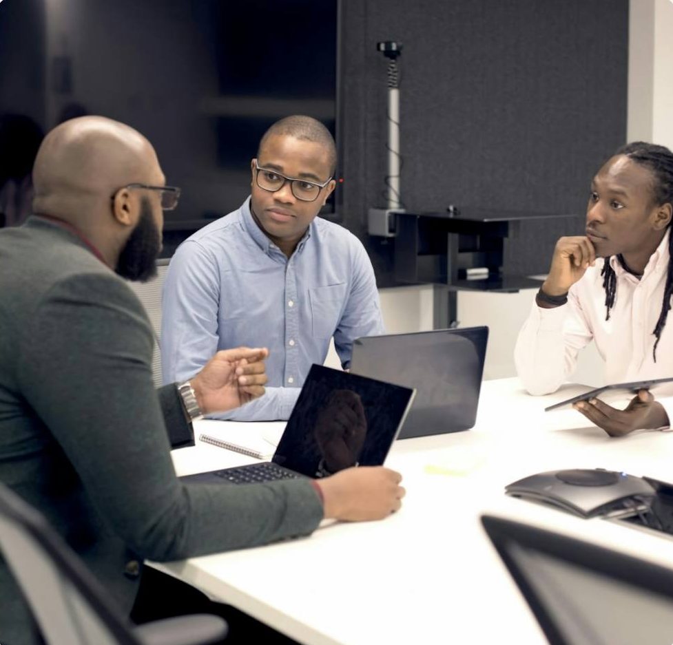 Three colleagues in discussion around a meeting table with laptops